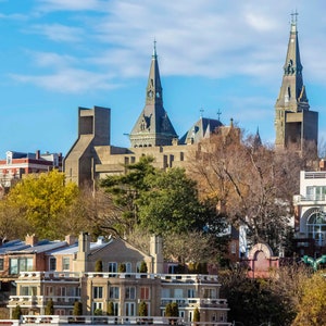 May include: A view of a city skyline with several buildings, including a tall building with a pointed roof and a smaller building with a flat roof. The buildings are surrounded by trees and bushes.