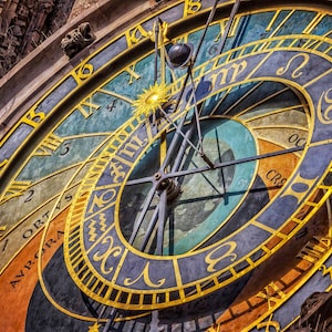 May include: Close-up of an ornate astronomical clock face with gold numerals and markings on a blue and teal background. The clock has a sun-like design in the center and is surrounded by architectural details.