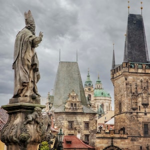 May include: A stone statue of a man in a robe stands on a pedestal with a cloudy sky and a tall stone tower in the background.