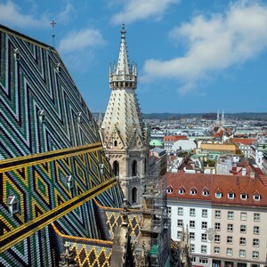 May include: A view of the city from the top of a church with a patterned roof. The church has a tall spire and is surrounded by buildings. The city is in the distance and the sky is blue with white clouds.
