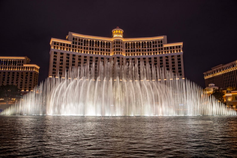 May include: A large, modern hotel with a facade of windows and a flat roof. The hotel is illuminated at night and has a large fountain in front of it. The fountain is shooting water high into the air, creating a dramatic display.