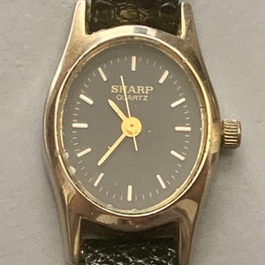May include: A silver-toned wristwatch with a black face and a black leather band. The watch face has white markings and the text "SHARP QUARTZ" in gold lettering.