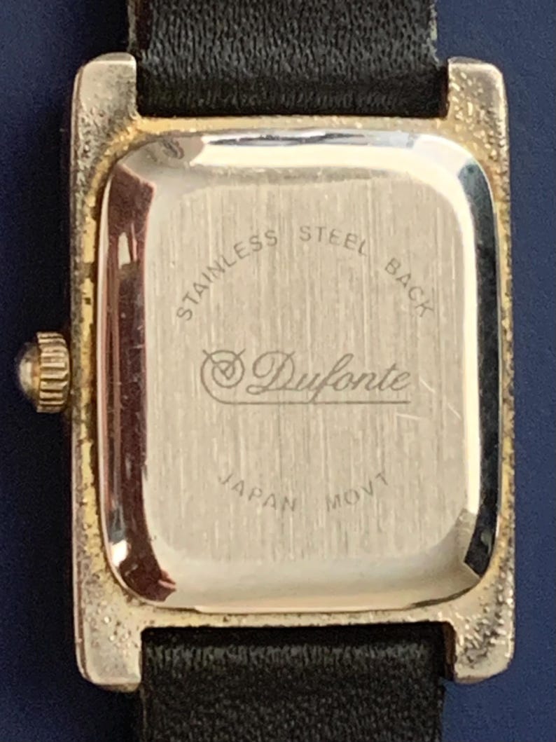 May include: The back of a gold-tone watch with a black leather band. The watch face is engraved with "Stainless Steel Back", "Duforte", and "Japan Movt".