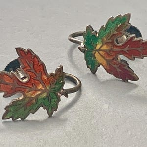 May include: A pair of vintage leaf-shaped earrings. Each earring features a detailed maple leaf design with a gradient of colors, including red, orange, yellow, and green. The earrings have a silver-tone metal ring and post.