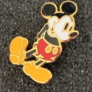 May include: A gold-colored Mickey Mouse pin. The pin features Mickey Mouse in his classic pose, with black ears, a white face, red shorts, and yellow shoes. The pin is set against a dark, textured background.