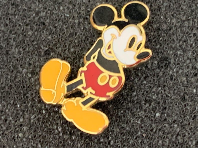 May include: A gold-colored Mickey Mouse pin. The pin features Mickey Mouse in his classic pose, with black ears, a white face, red shorts, and yellow shoes. The pin is set against a dark, textured background.