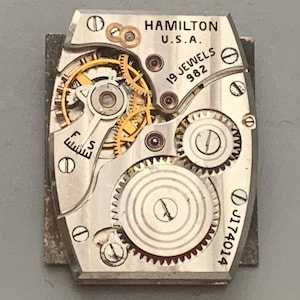 May include: A vintage Hamilton watch movement, featuring intricate gears and mechanisms. The silver-toned plate is marked "HAMILTON U.S.A." and "19 JEWELS 982." The serial number "J174014" is also visible. The watch movement is a rectangular shape.