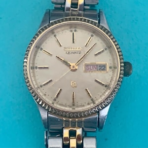 May include: A two-tone Citizen Quartz wristwatch with a silver and gold-colored metal link band. The round watch face features a light-colored dial with gold-tone hour markers, gold hands, and a day-date window displaying SUN 22 at the three o'clock position.
