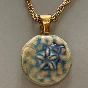 May include: A gold-toned necklace with a round pendant. The pendant features a blue and white floral design. The chain is a twisted rope style.