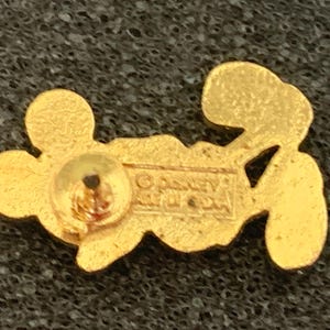 May include: A gold-colored pin featuring a stylized Mickey Mouse design. The pin has a post and clasp on the back for attachment. The back of the pin has the text "&copy; Disney" and other text. The pin is on a textured, dark surface.