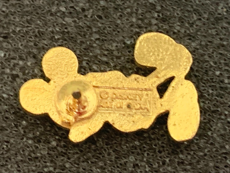 May include: A gold-colored pin featuring a stylized Mickey Mouse design. The pin has a post and clasp on the back for attachment. The back of the pin has the text "&copy; Disney" and other text. The pin is on a textured, dark surface.