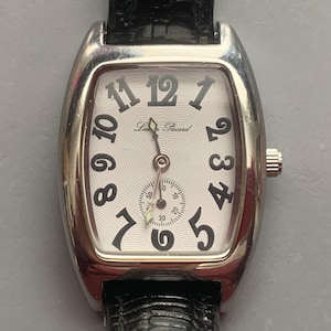 May include: A silver-toned wristwatch with a rectangular case and a white dial featuring a wavy texture. The dial displays black Arabic numerals, the text Lucien Piccard, and a small sub-dial for seconds. It has a black textured leather strap.