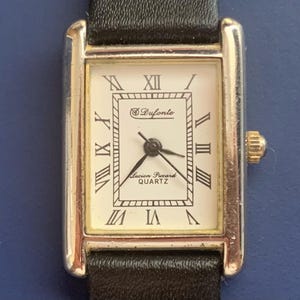 May include: A silver-toned wristwatch with a white face and black leather band. The watch face has black Roman numeral hour markers and a small seconds dial. The watch face also has the text "Drufonte" and "Lucien Piccard Quartz".