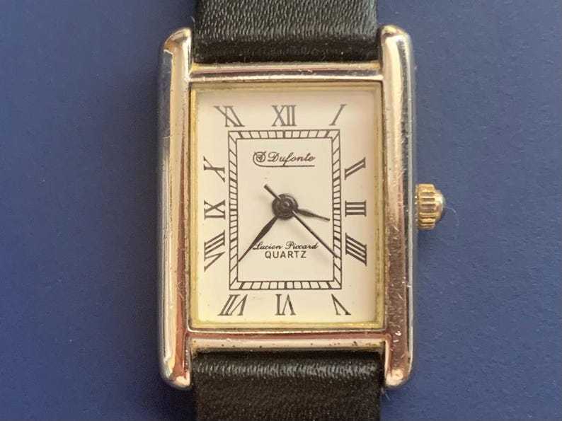 May include: A silver-toned wristwatch with a white face and black leather band. The watch face has black Roman numeral hour markers and a small seconds dial. The watch face also has the text "Drufonte" and "Lucien Piccard Quartz".