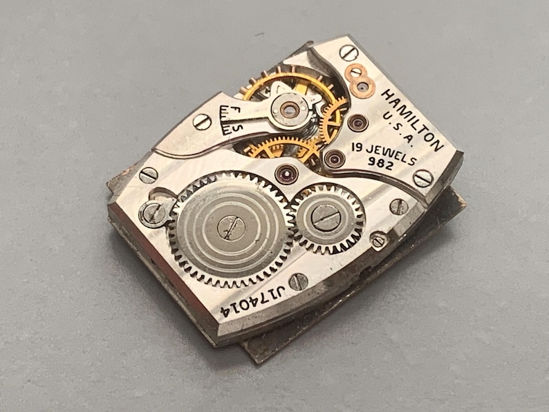 May include: Close-up of a vintage Hamilton watch movement. The silver-toned mechanism features intricate gears, jewels, and the text "HAMILTON U.S.A. 19 JEWELS 982". The serial number "J174014" is also visible.