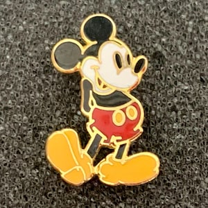May include: A gold-toned Mickey Mouse pin. The pin features Mickey in his classic pose, with a black head and ears, white face, black torso, red shorts, and yellow shoes. The pin has a gold border.