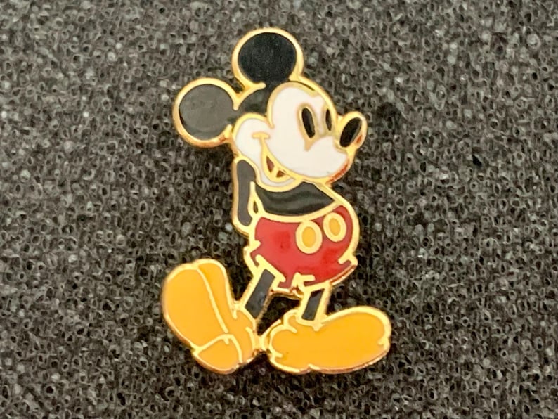 May include: A gold-toned Mickey Mouse pin. The pin features Mickey in his classic pose, with a black head and ears, white face, black torso, red shorts, and yellow shoes. The pin has a gold border.