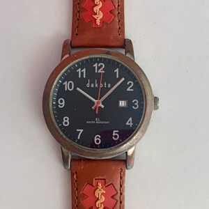 May include: A Dakota wristwatch with a black face, white numerals, and a date window at the three o'clock position. The watch features a brown leather strap adorned with red medical alert symbols featuring a gold-colored caduceus emblem.