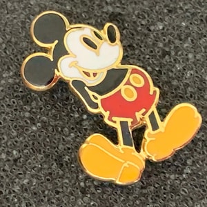 May include: A gold-colored Mickey Mouse enamel pin. The pin features Mickey Mouse in his classic pose, with black ears, a white face, red shorts, and yellow shoes. The pin has a gold border and is set against a textured gray background.