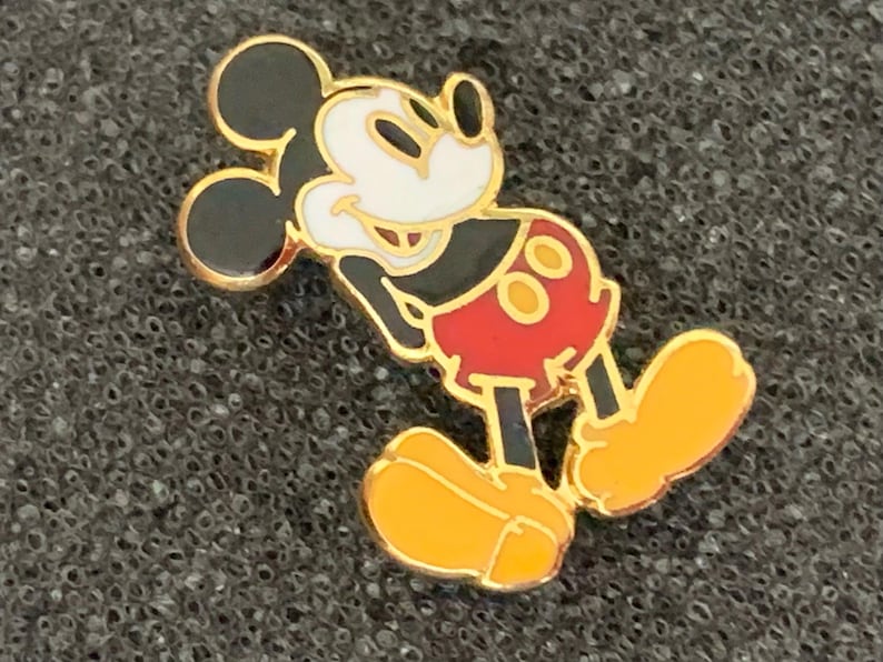 May include: A gold-colored Mickey Mouse enamel pin. The pin features Mickey Mouse in his classic pose, with black ears, a white face, red shorts, and yellow shoes. The pin has a gold border and is set against a textured gray background.