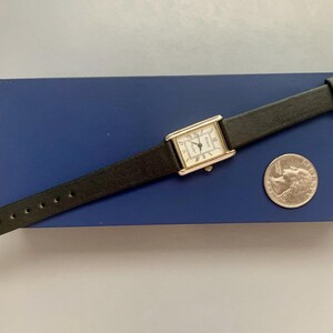 May include: A silver watch with a white face and black leather band. The watch is on a blue background with a quarter next to it for scale.
