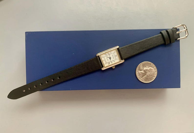 May include: A silver watch with a white face and black leather band. The watch is on a blue background with a quarter next to it for scale.
