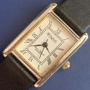 May include: A silver and gold rectangular watch with a white face and black leather band. The watch face has Roman numeral hour markers and the text "Swiss Quartz" and "S. Dufonte".