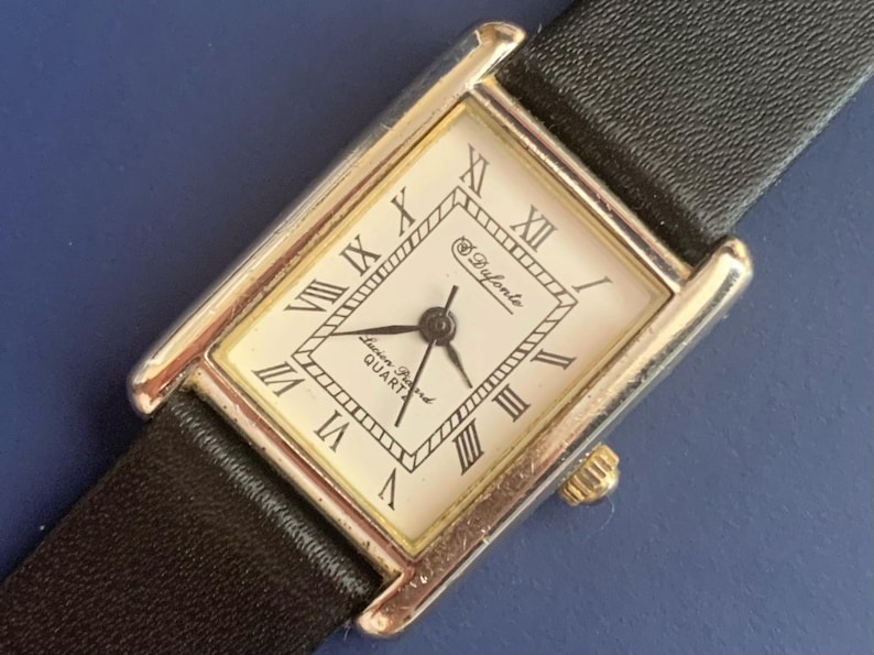 May include: A silver and gold rectangular watch with a white face and black leather band. The watch face has Roman numeral hour markers and the text "Swiss Quartz" and "S. Dufonte".