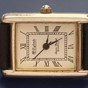May include: A silver-toned square wristwatch with a white face and black Roman numeral hour markers. The watch has a black leather band and says "S. Dyferente" and "Swiss Quartz" on the face.