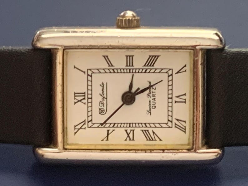 May include: A silver-toned square wristwatch with a white face and black Roman numeral hour markers. The watch has a black leather band and says "S. Dyferente" and "Swiss Quartz" on the face.