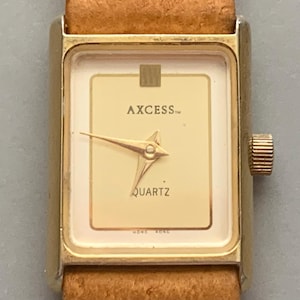 May include: A gold-toned AXCESS quartz watch with a rectangular face and a light brown leather band. The watch face is cream-colored with gold hands and the word "QUARTZ" printed on it. The watch is a classic style.