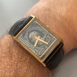 May include: A rectangular gold-tone wristwatch featuring a black United States postage stamp on the dial with a portrait of George Washington. The watch has a black leather strap with a textured finish and a small gold-tone crown on the side.