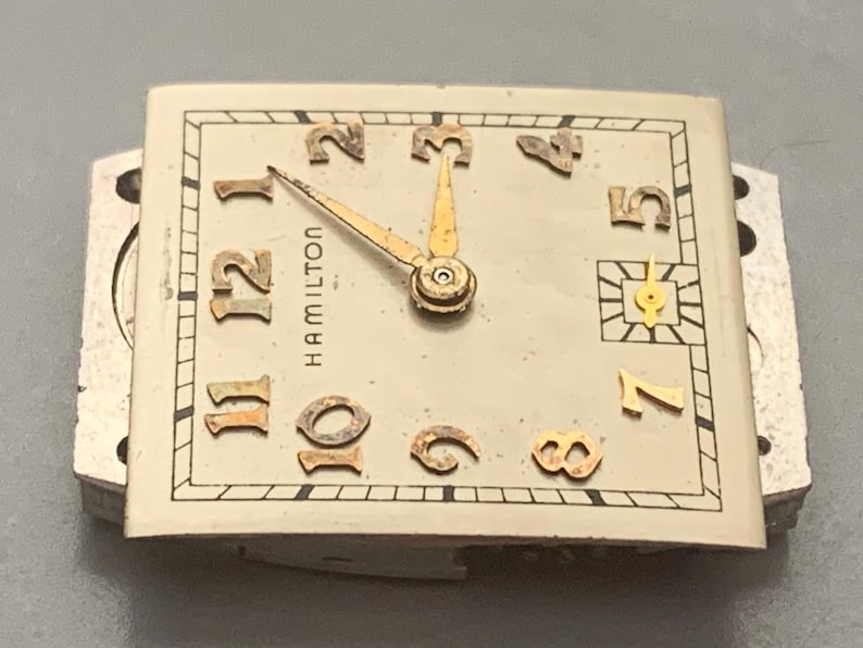 May include: Vintage Hamilton watch face with gold-tone hands and numerals on a cream-colored background. The watch face is rectangular with the brand name "HAMILTON" printed vertically. The numerals are in a classic font.