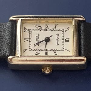 May include: A gold-toned wristwatch with a black leather band. The watch face is white with black Roman numeral hour markers and the text "Lauren Ralph Lauren QUARTZ" and "&copy; Bulova" printed on it.