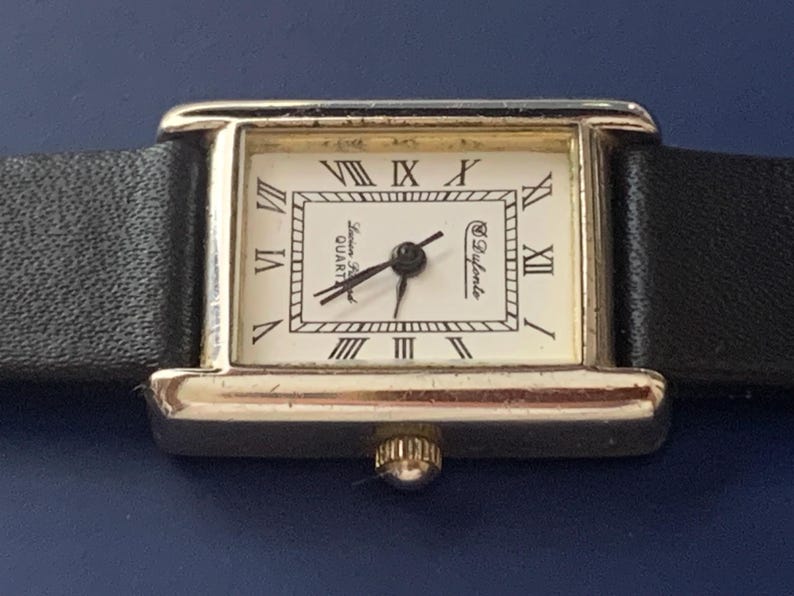 May include: A gold-toned wristwatch with a black leather band. The watch face is white with black Roman numeral hour markers and the text "Lauren Ralph Lauren QUARTZ" and "&copy; Bulova" printed on it.
