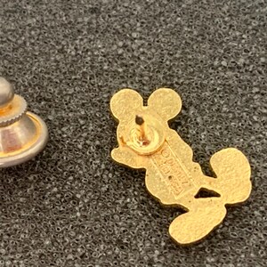 May include: A gold-colored Mickey Mouse lapel pin with a matching pin back. The pin is in the shape of Mickey Mouse and is lying on a textured, dark gray surface. The pin back is a separate piece.