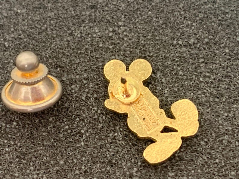 May include: A gold-colored Mickey Mouse lapel pin with a matching pin back. The pin is in the shape of Mickey Mouse and is lying on a textured, dark gray surface. The pin back is a separate piece.