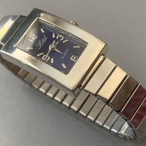 May include: Silver-toned quartz watch with a rectangular face and a dark blue dial. The watch features the text "Ltd." and "Quartz". The watch band is a silver-toned, expandable style.