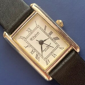 May include: A silver and gold square-shaped wristwatch with a black leather band. The watch face has Roman numeral hour markers and the text "Swiss Ryfonte Precision Quartz".