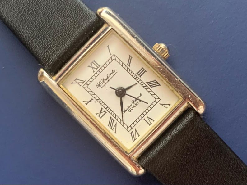 May include: A silver and gold square-shaped wristwatch with a black leather band. The watch face has Roman numeral hour markers and the text "Swiss Ryfonte Precision Quartz".