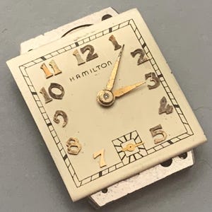 May include: Vintage Hamilton watch face with a cream-colored background. The watch face features gold-colored numerals and hands, with the brand name "HAMILTON" displayed. The watch hands point to approximately 3:10. The watch face is rectangular.