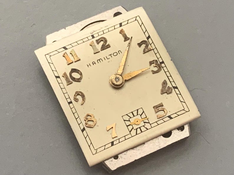 May include: Vintage Hamilton watch face with a cream-colored background. The watch face features gold-colored numerals and hands, with the brand name "HAMILTON" displayed. The watch hands point to approximately 3:10. The watch face is rectangular.
