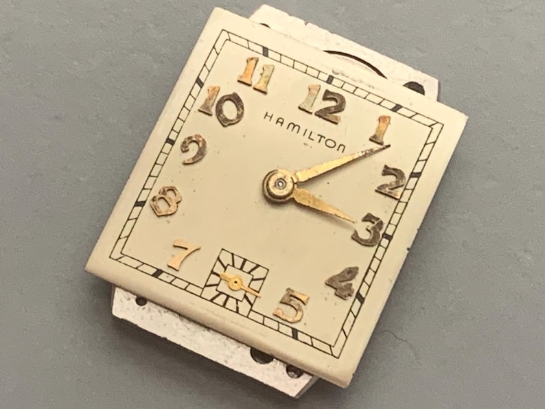 May include: Vintage Hamilton watch face with a rectangular shape. The off-white face features gold-colored numerals and hands. The word "HAMILTON" is printed in black. The watch face is set against a gray background.