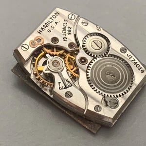 May include: Close-up of a vintage Hamilton watch movement. The silver-toned mechanism features intricate gears, jewels, and the text "HAMILTON U.S.A. 19 JEWELS 982" and "J174014". The watch movement is rectangular and has a complex internal structure.
