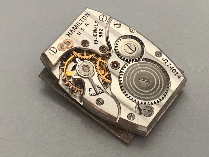 May include: Close-up of a vintage Hamilton watch movement. The silver-toned mechanism features intricate gears, jewels, and the text "HAMILTON U.S.A. 19 JEWELS 982" and "J174014". The watch movement is rectangular and has a complex internal structure.