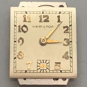 May include: Vintage Hamilton watch face with a rectangular shape. The watch face is cream-colored with gold-toned numerals and hands. The word "HAMILTON" is printed in black. The watch hands point to 2:15.