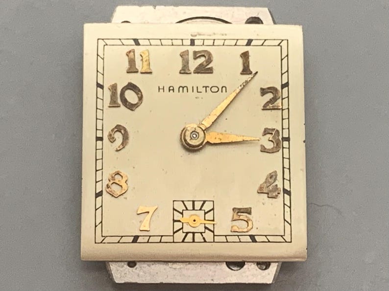 May include: Vintage Hamilton watch face with a rectangular shape. The watch face is cream-colored with gold-toned numerals and hands. The word "HAMILTON" is printed in black. The watch hands point to 2:15.