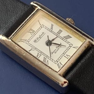 May include: A silver-toned rectangular watch with a white face and black leather band. The watch face has Roman numeral hour markers and the text "Swiss Made Quartz" printed on it. The watch brand is "D'Avorio".