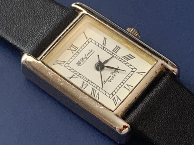 May include: A silver-toned rectangular watch with a white face and black leather band. The watch face has Roman numeral hour markers and the text "Swiss Made Quartz" printed on it. The watch brand is "D'Avorio".