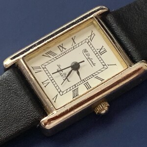 May include: A gold-tone rectangular watch with a white face and black leather band. The watch face has Roman numeral hour markers and the text "Le D'yante Quartz" printed on it.